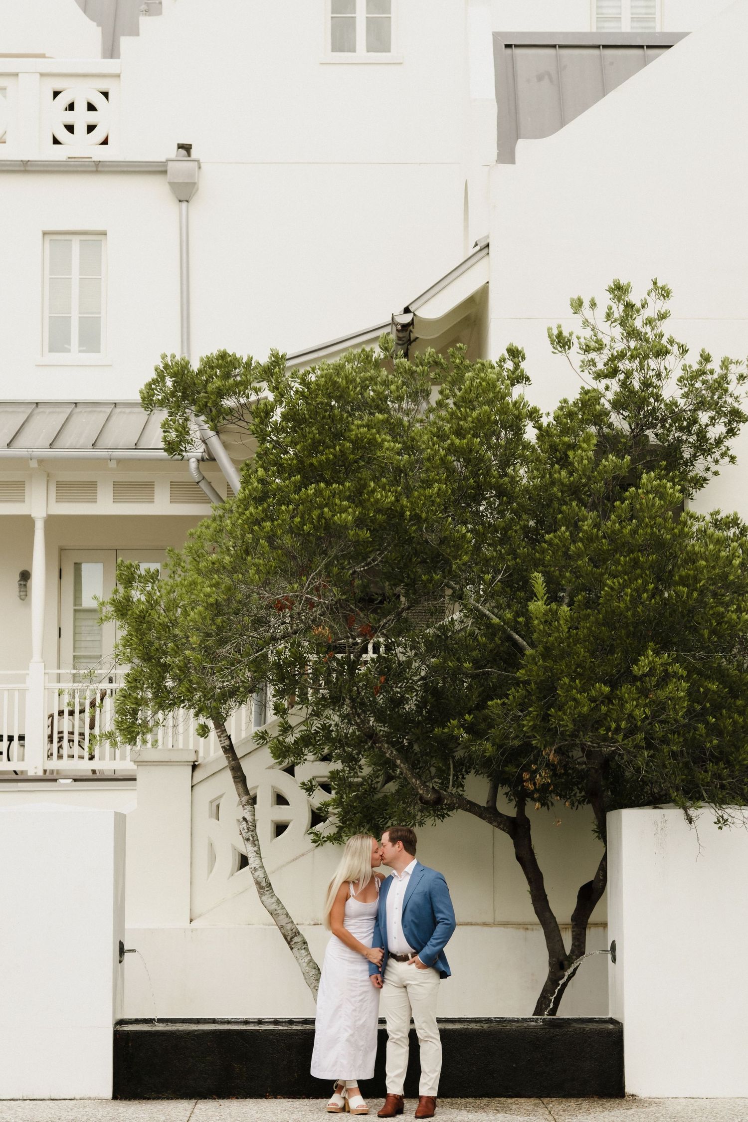 Couple poses together against white architectural backdrop with trees.