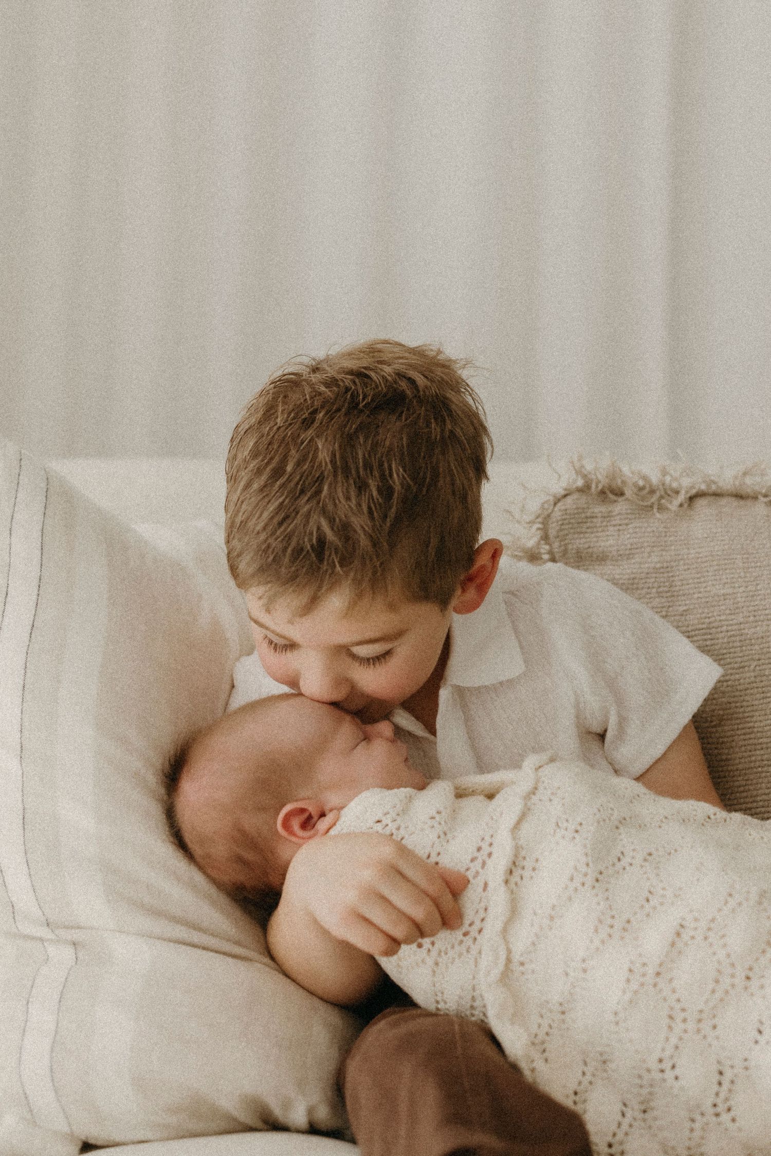 Young boy gently kissing newborn baby sibling wrapped in knit blanket on sofa.