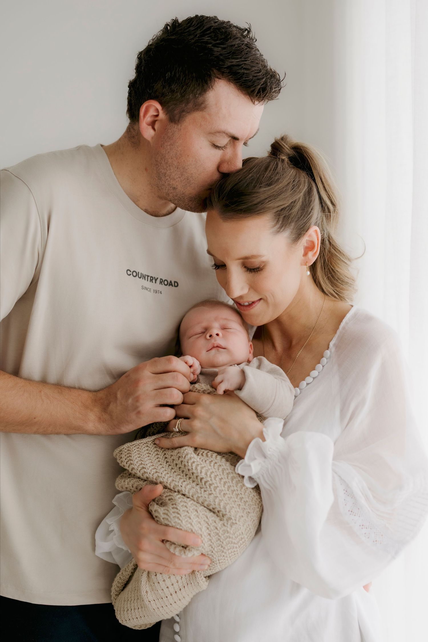 New parents in neutral tones tenderly holding and kissing their sleeping newborn baby wrapped in knit blanket.