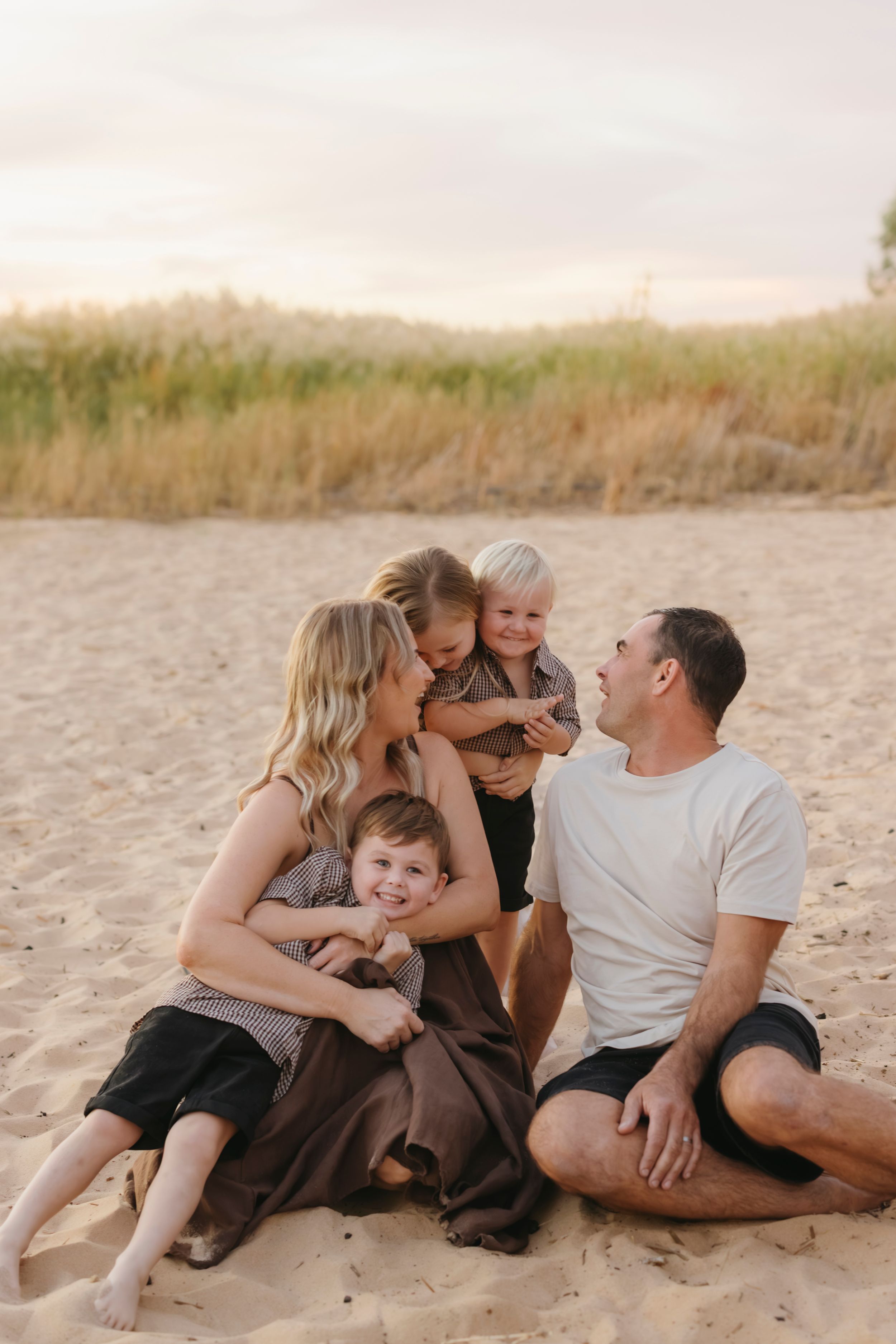 A happy family of five sits together on a sandy beach at sunset, laughing and embracing.