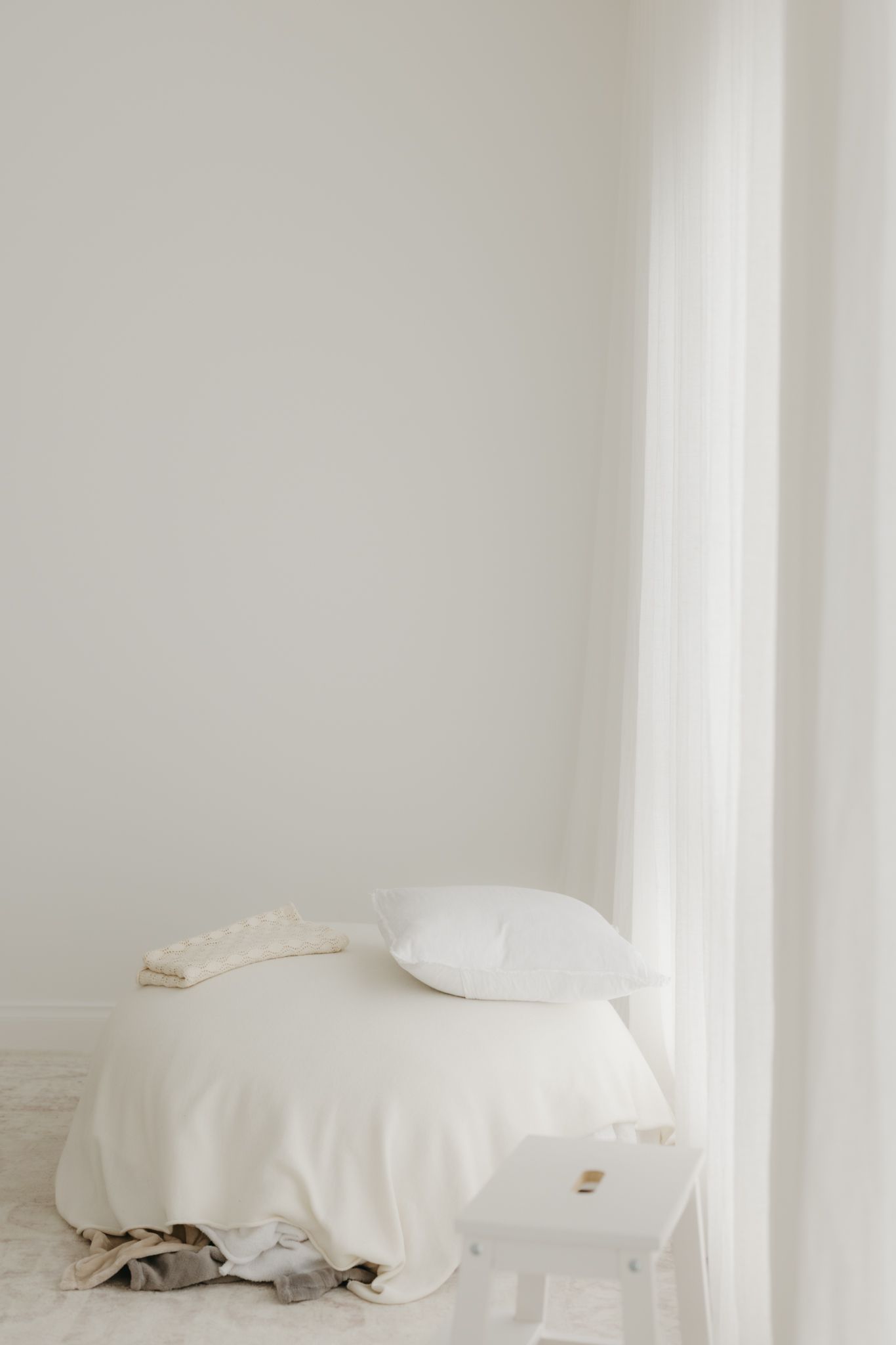 Minimalist white bedroom corner with a neatly made bed, white pillow, and sheer curtains by a window.