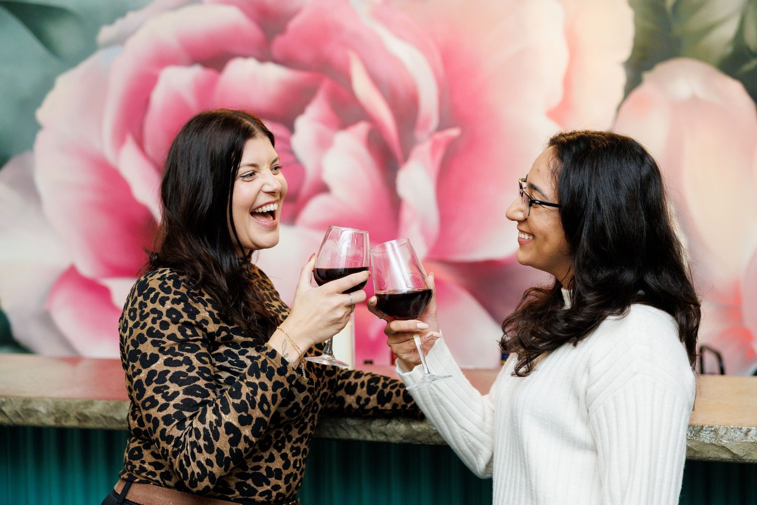 Two friends enjoy wine and conversation together against a pink floral backdrop in a series of candid moments.