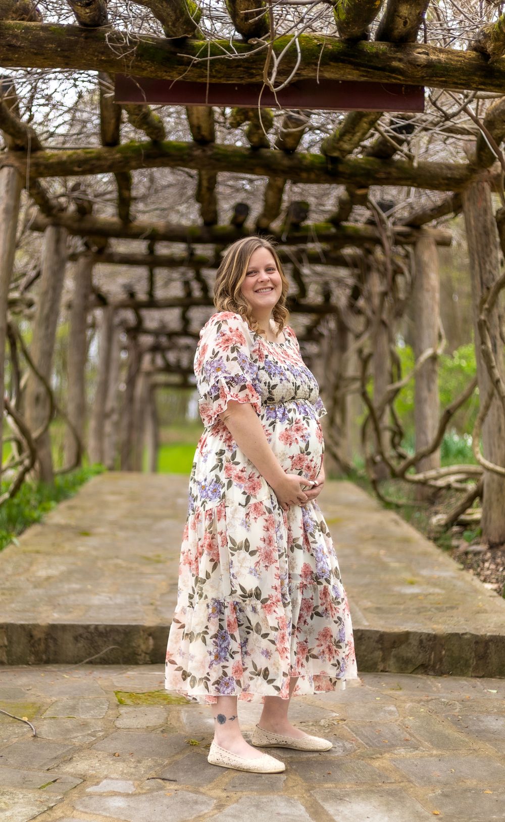 A floral maternity photoshoot under a rustic wooden pergola with vines.
