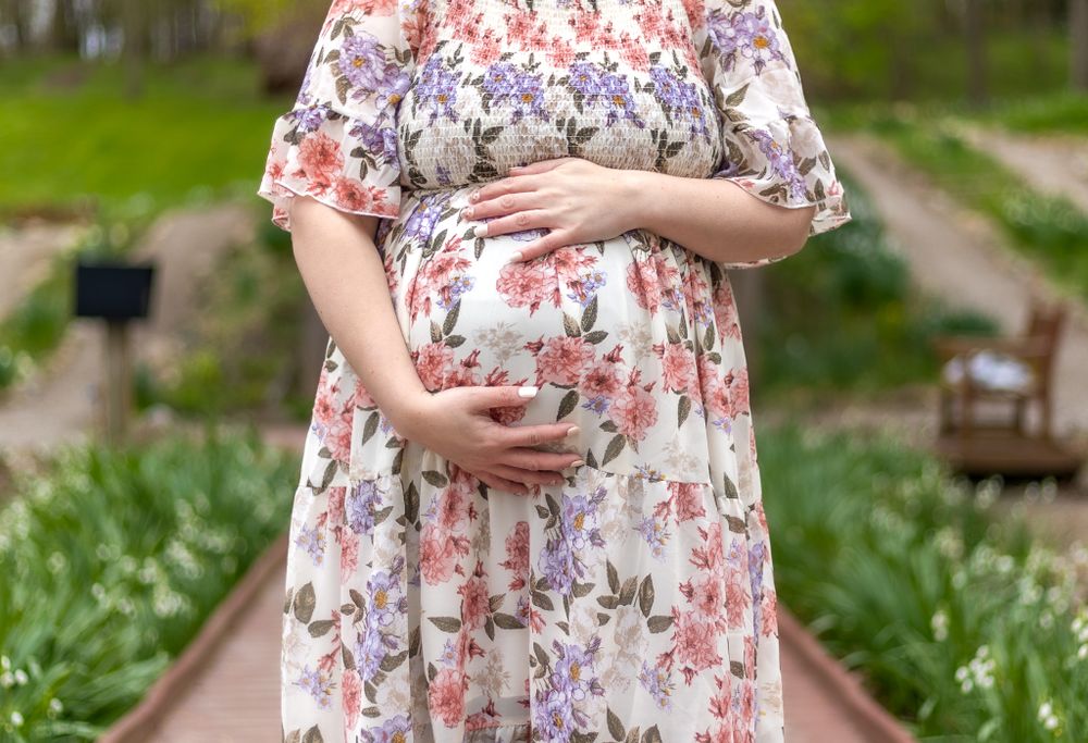 Close-up of hands cradling a pregnant belly in a floral dress against a garden background.