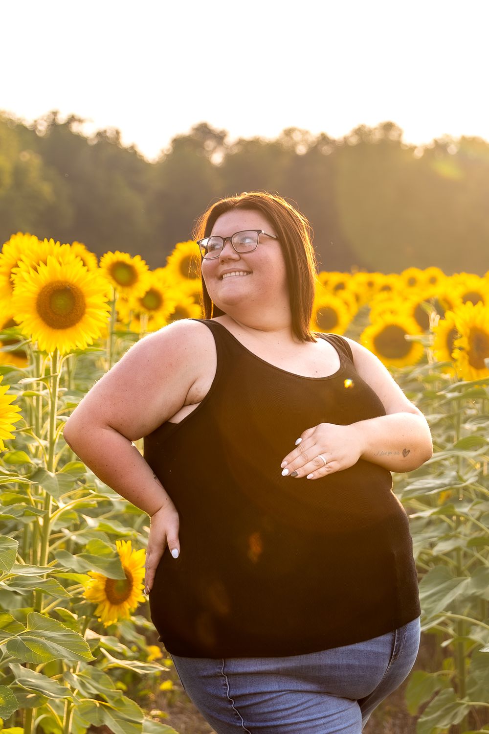 Woman in black tank top poses joyfully in vibrant sunflower field at sunset.