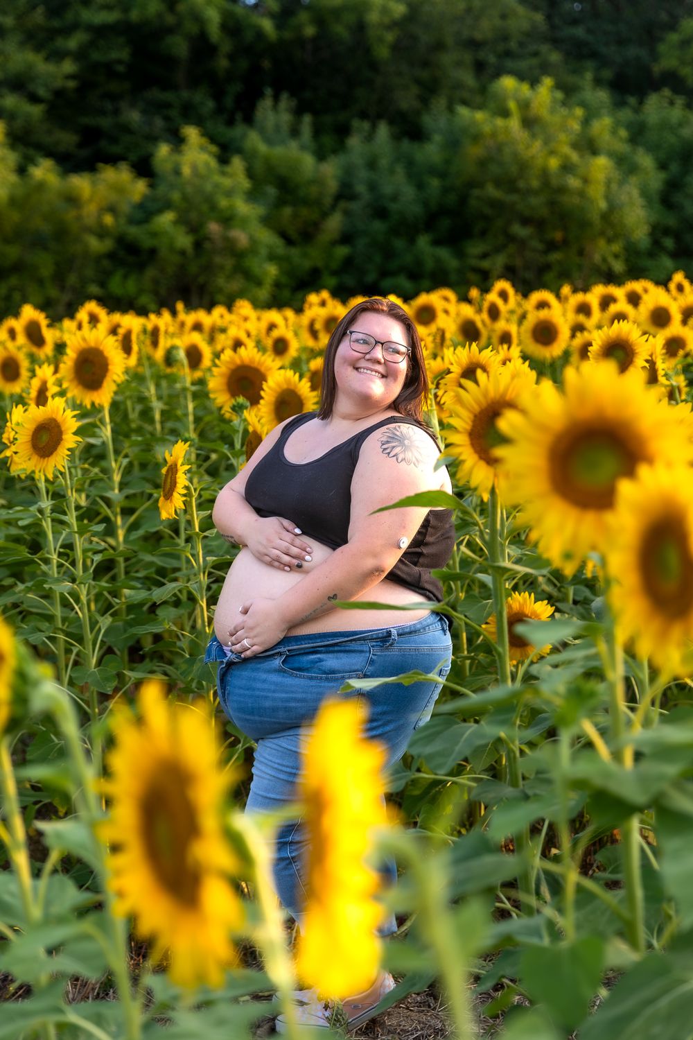 Woman in dark top stands confidently among blooming sunflowers in golden sunlight.