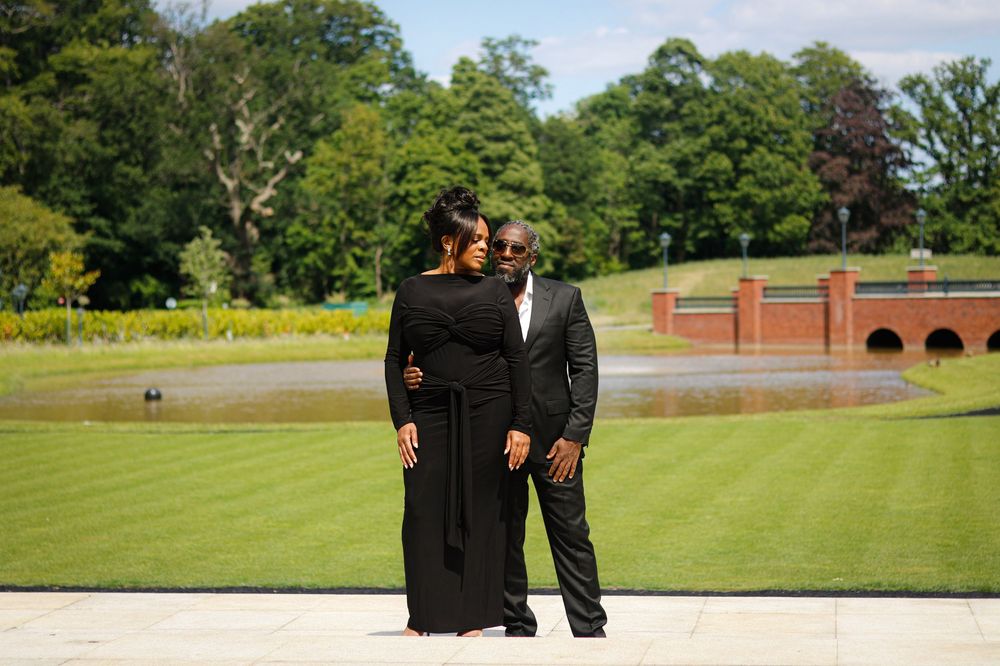 Couple in formal attire walking on manicured lawn with brick building in background.