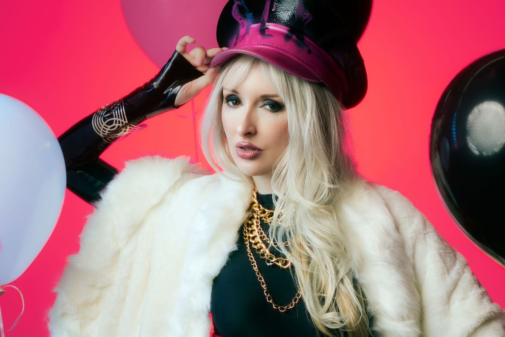 Blonde woman wearing a black outfit, white faux fur shawl, gold chains and a pink pilot hat, posing against a vibrant pink backdrop balloons