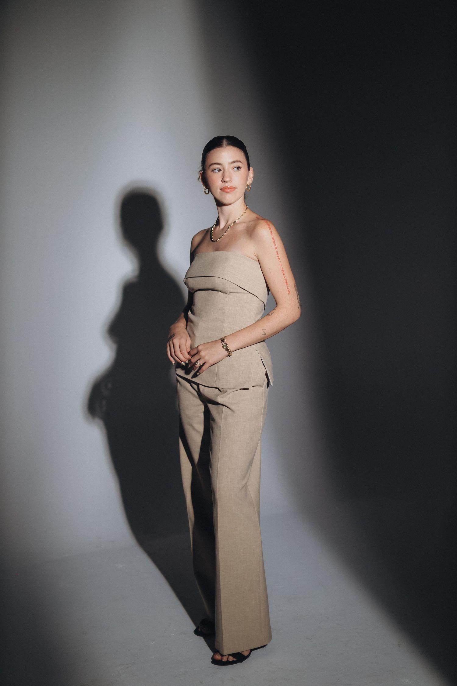 Fashion model in beige outfit poses against white wall creating dramatic shadow effect.