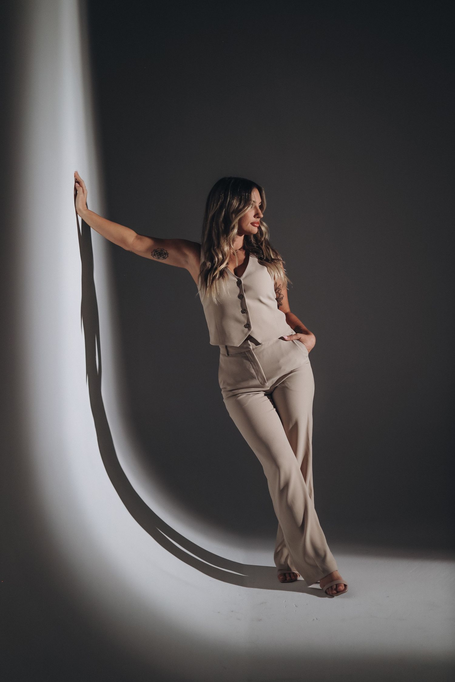 Professional model in beige outfit poses dramatically against curved light beam in artistic studio shot.