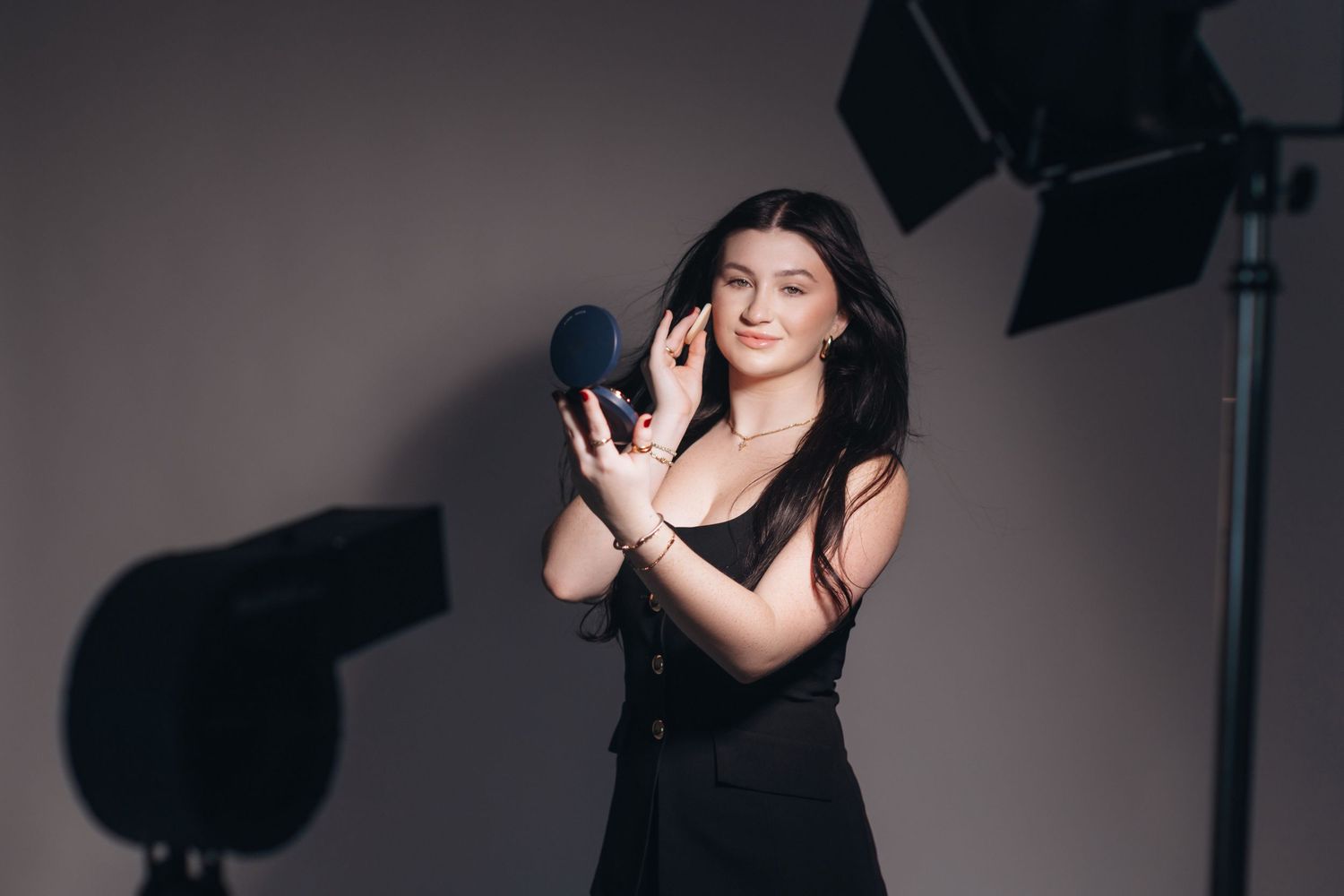 A person in a black dress gestures with hands during a professional photoshoot with studio lights visible.