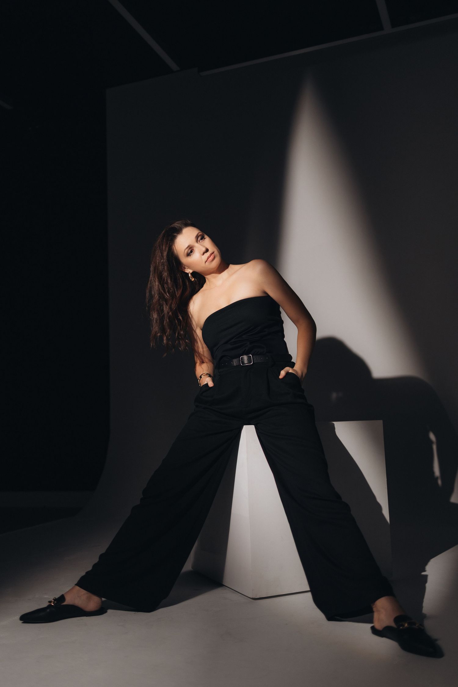 Fashion model in black wide-leg pants poses against white cube in dramatic lighting.
