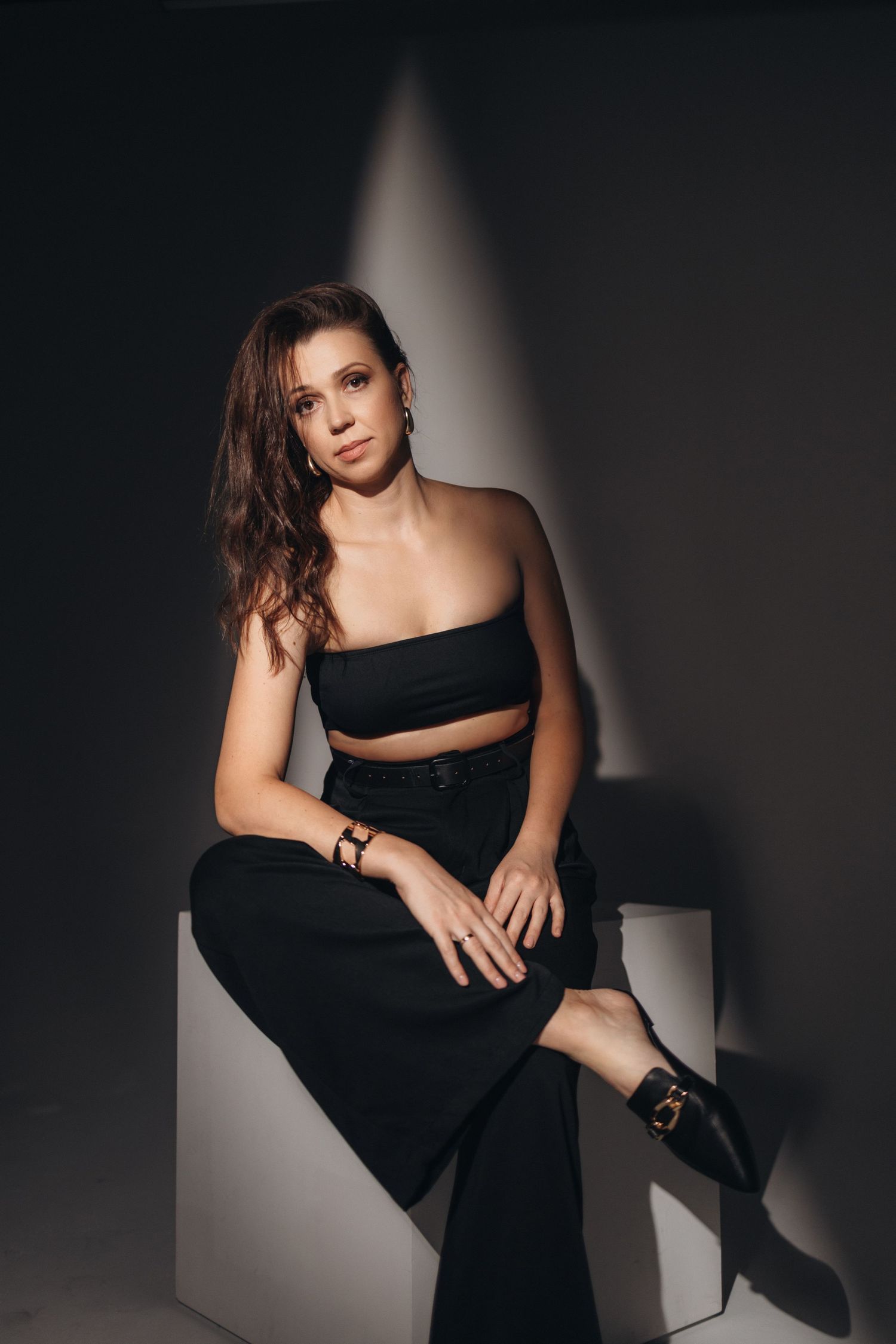 Modern portrait of a person in black outfit sitting on white cube with moody lighting.