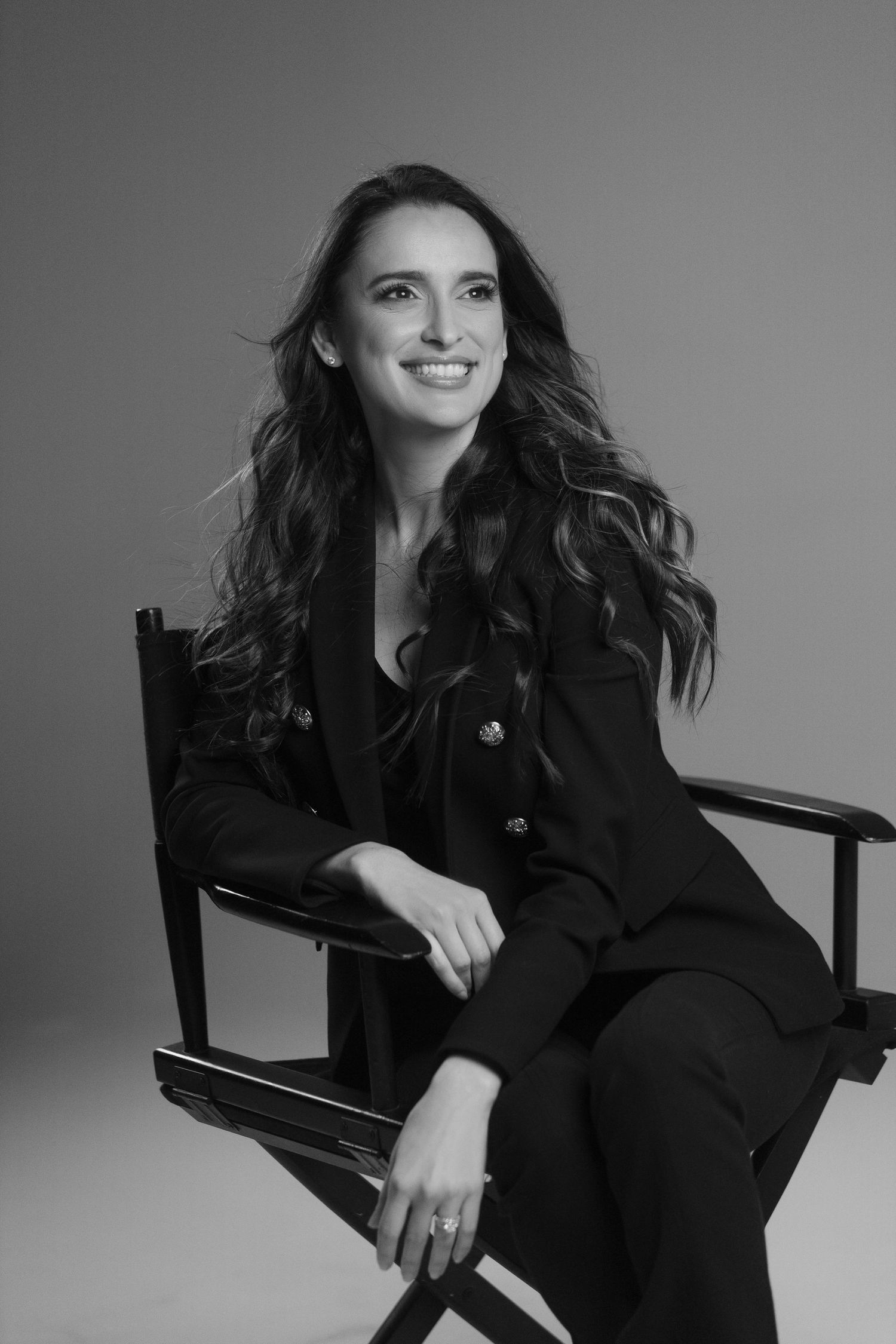 Black and white studio portrait of someone with long dark hair seated in a director's chair wearing a dark suit.