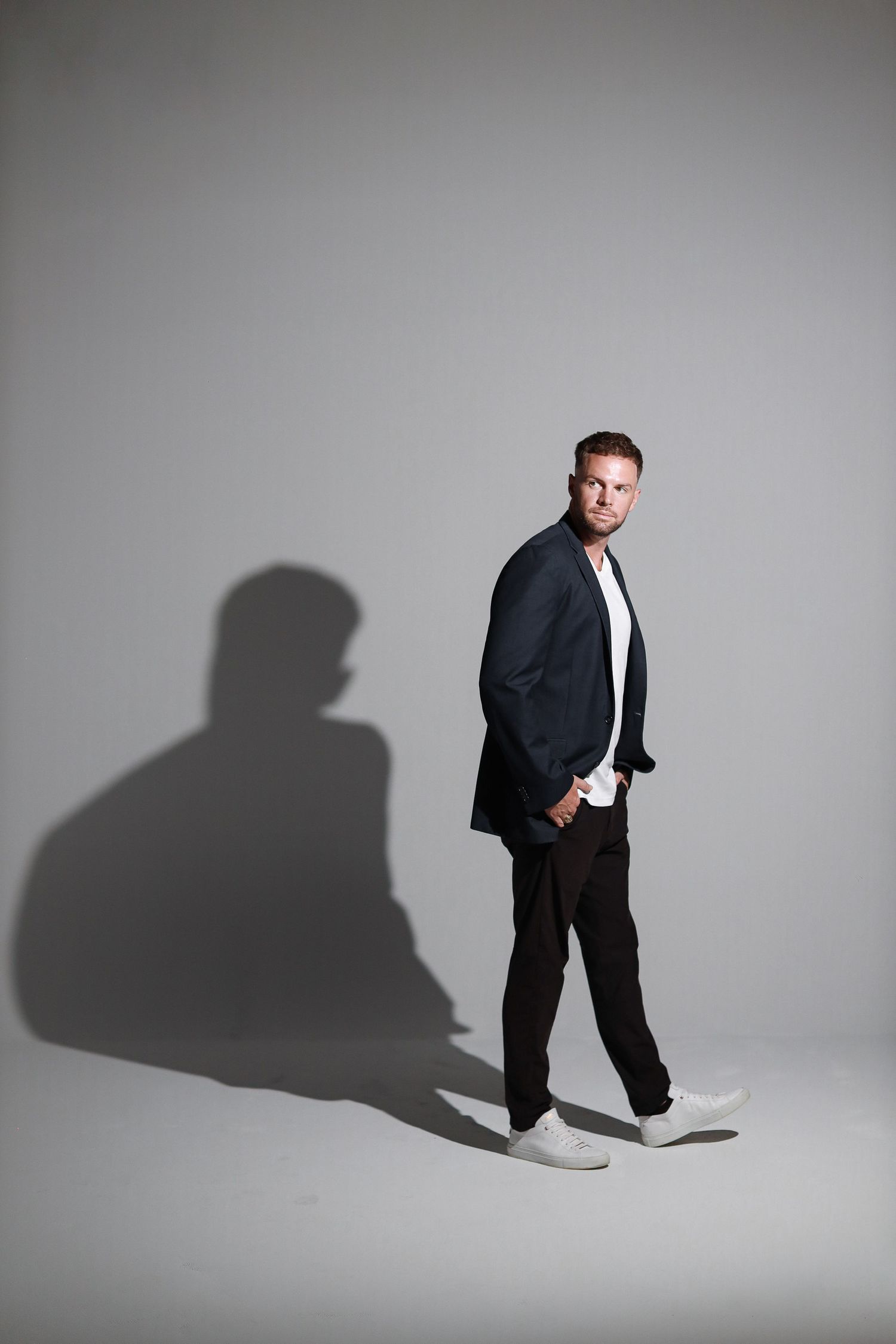 Fashion portrait of man in dark blazer with dramatic shadow cast on light studio background.