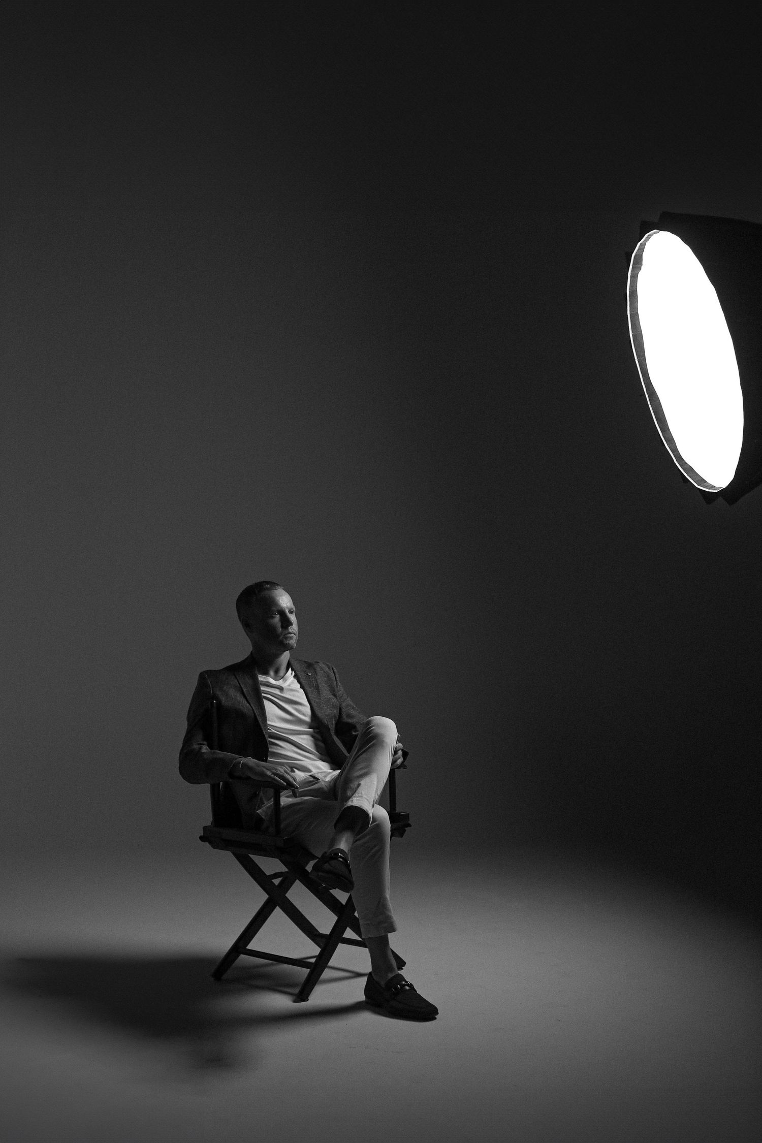 Dramatic studio lighting illuminates seated figure in dark setting with spotlight visible.