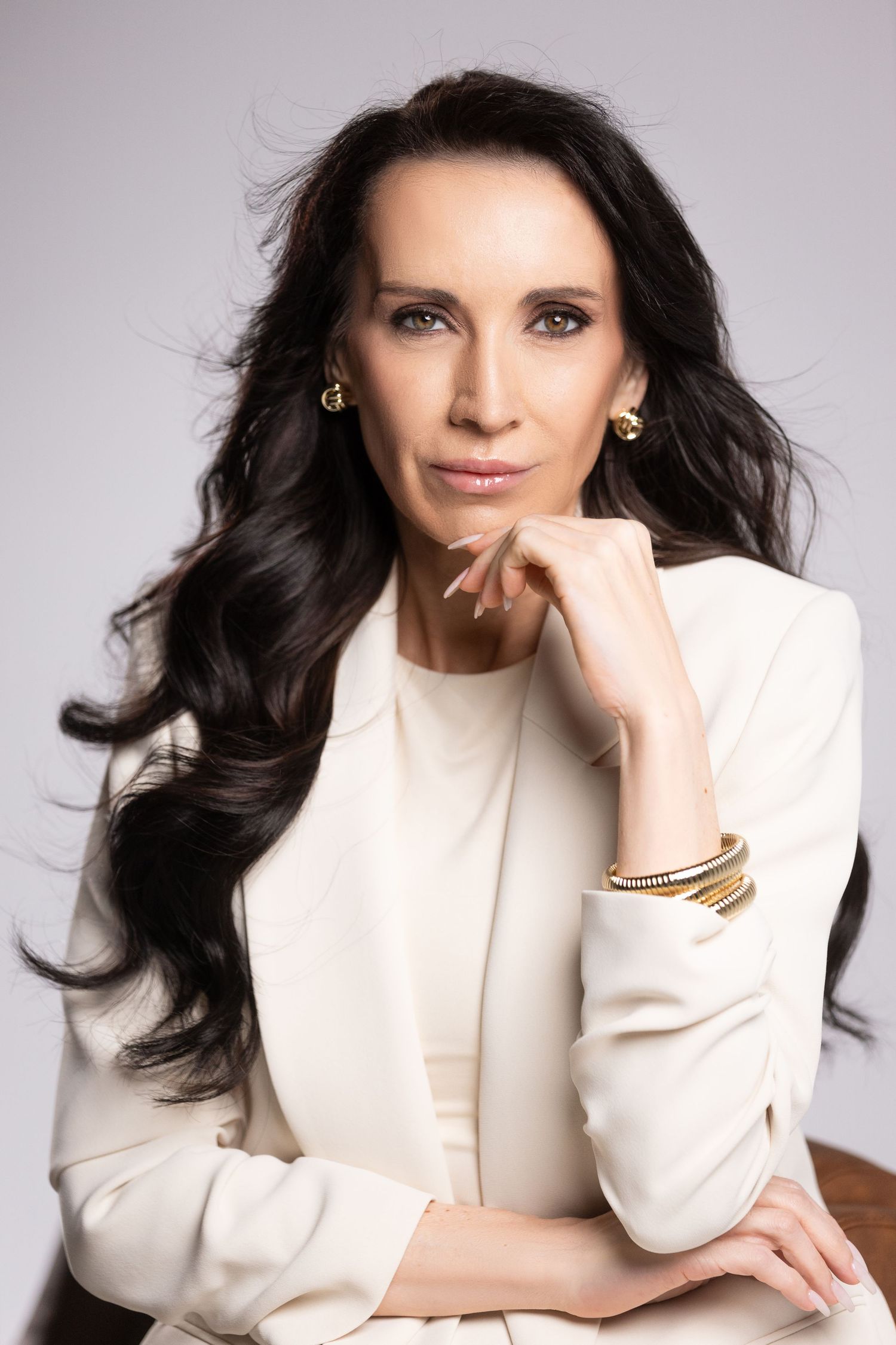 Professional portrait of a person with long dark wavy hair wearing a white blazer and gold bracelets against a light background.
