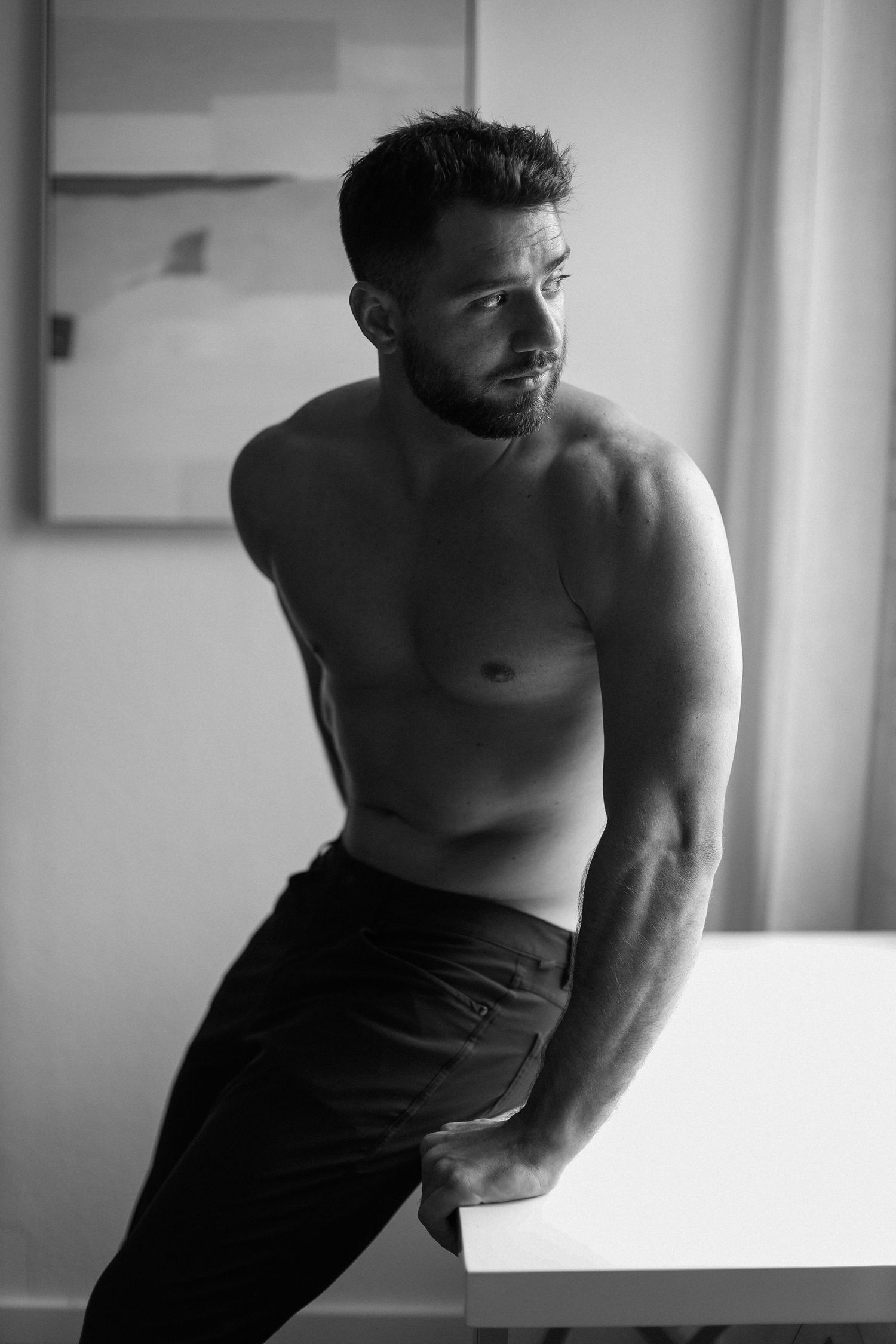 Artistic black and white portrait of a shirtless muscular man leaning against a counter.