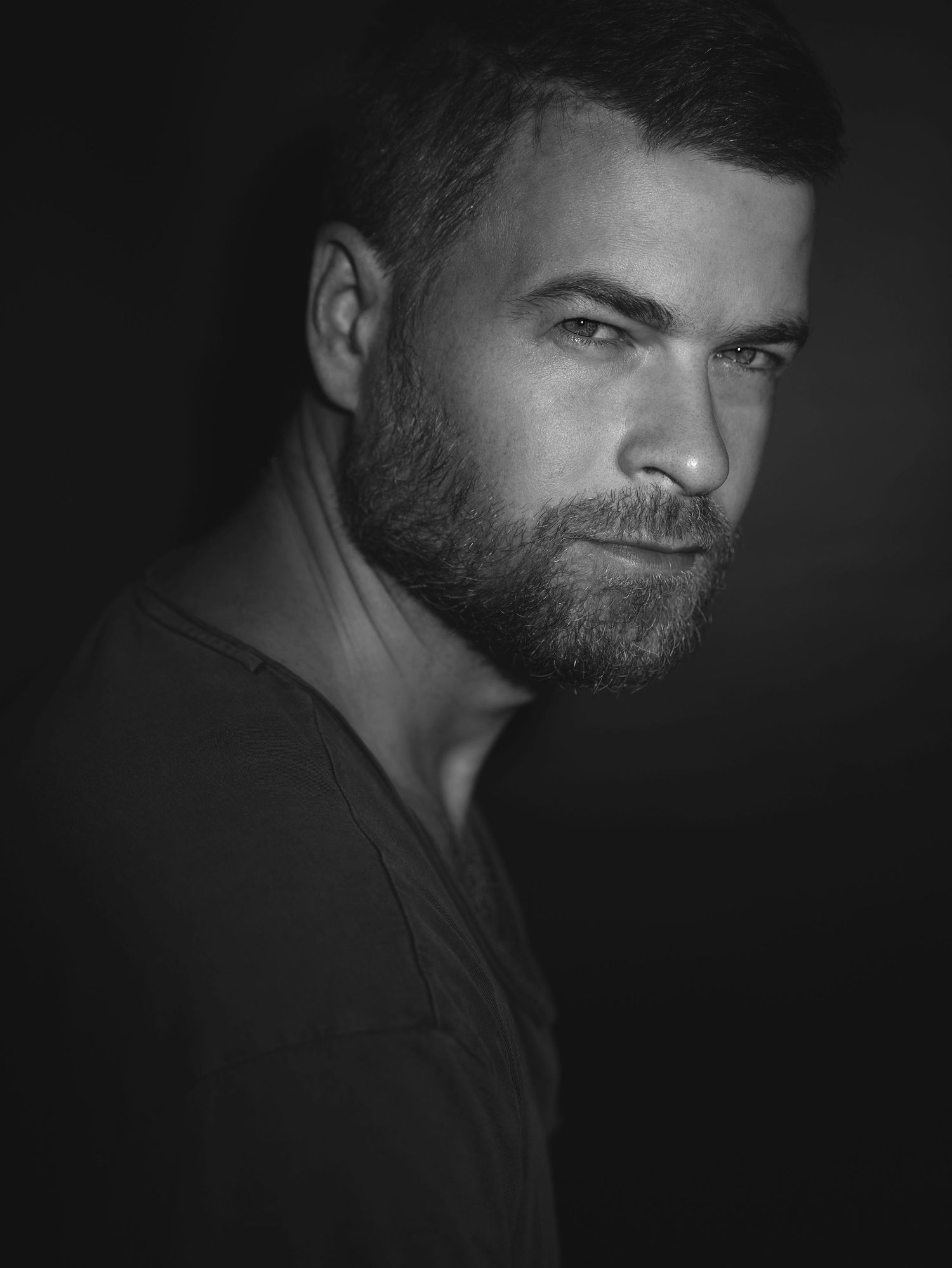 Moody black and white close-up portrait of a bearded man in dark lighting.