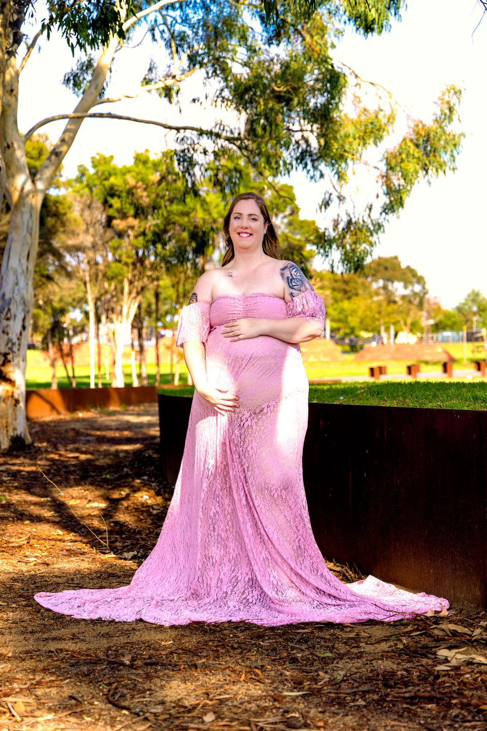 Pregnant woman in pink lace maternity gown posing outdoors in a park surrounded by eucalyptus trees.