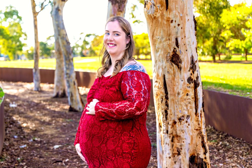 Pregnant woman in red lace dress posing by a tree in a park, smiling warmly during maternity photoshoot.