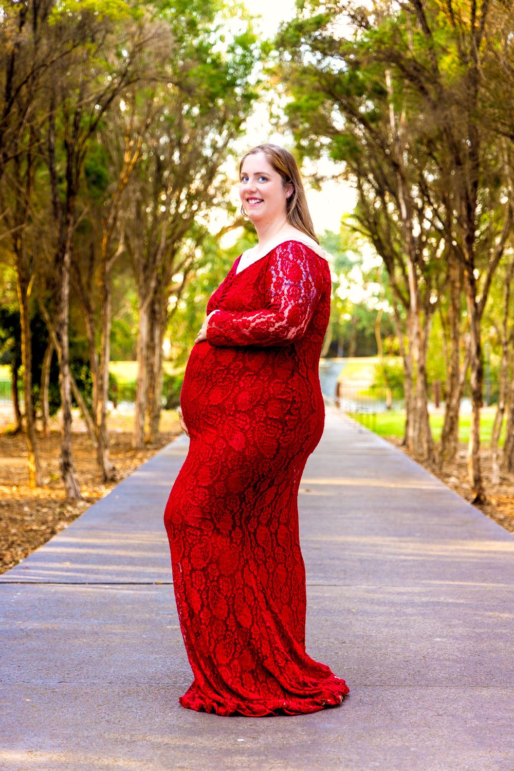Pregnant woman in red lace gown posing on a tree-lined path in a park during a maternity photo shoot.