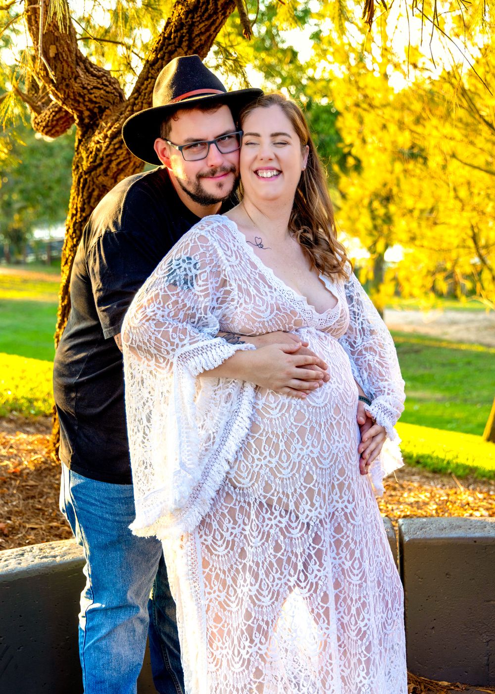 Couple posing outdoors for maternity photo, man in black hat embracing pregnant woman in white lace dress, autumn trees background.