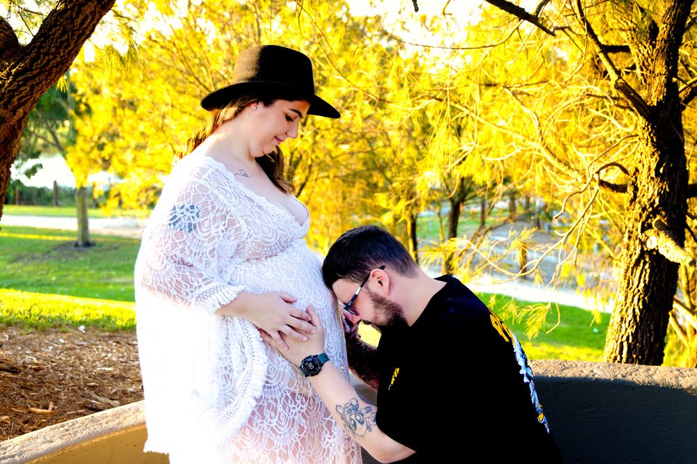 Pregnant woman in white lace dress and black hat with partner kneeling beside her in autumn park.