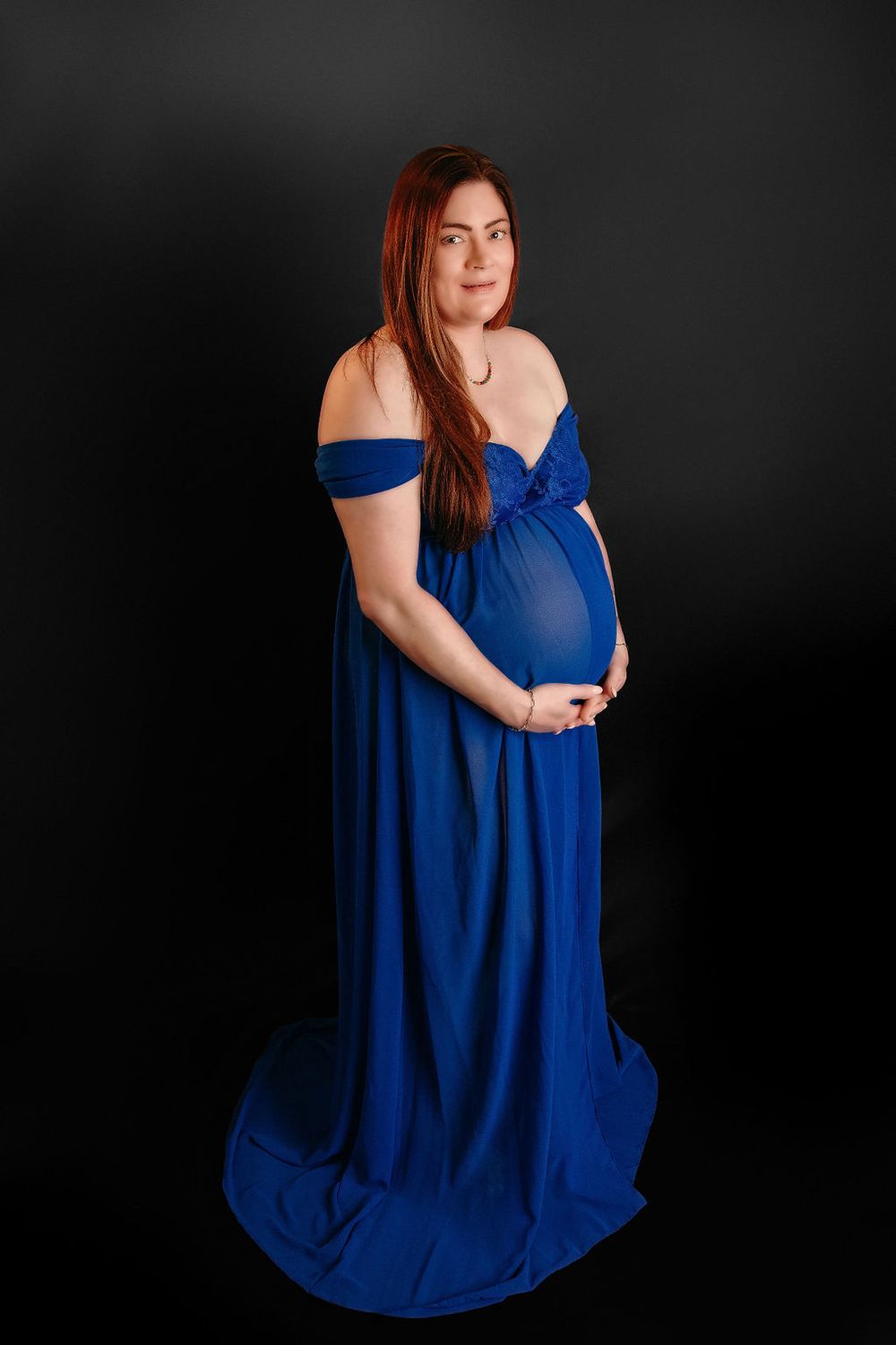 Full-length maternity portrait in royal blue gown against dark backdrop.