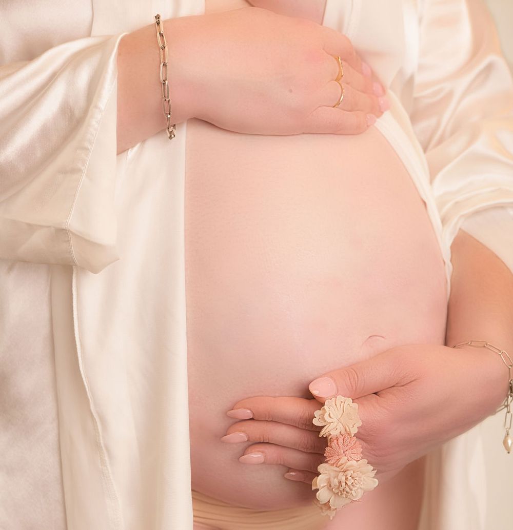 Close-up detail shot of pregnant belly with delicate floral accessories and white silk fabric.