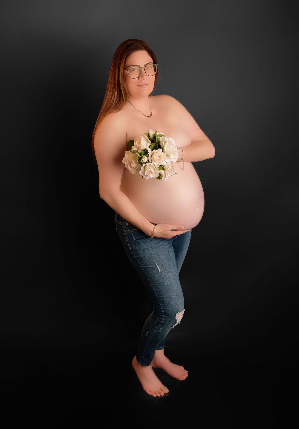 Maternity photo of expectant parent in jeans holding white roses against dark background.
