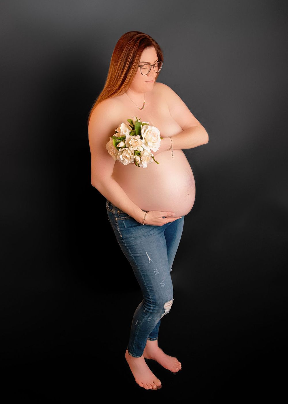 Side view of pregnant person in glasses holding white flowers against dark backdrop.