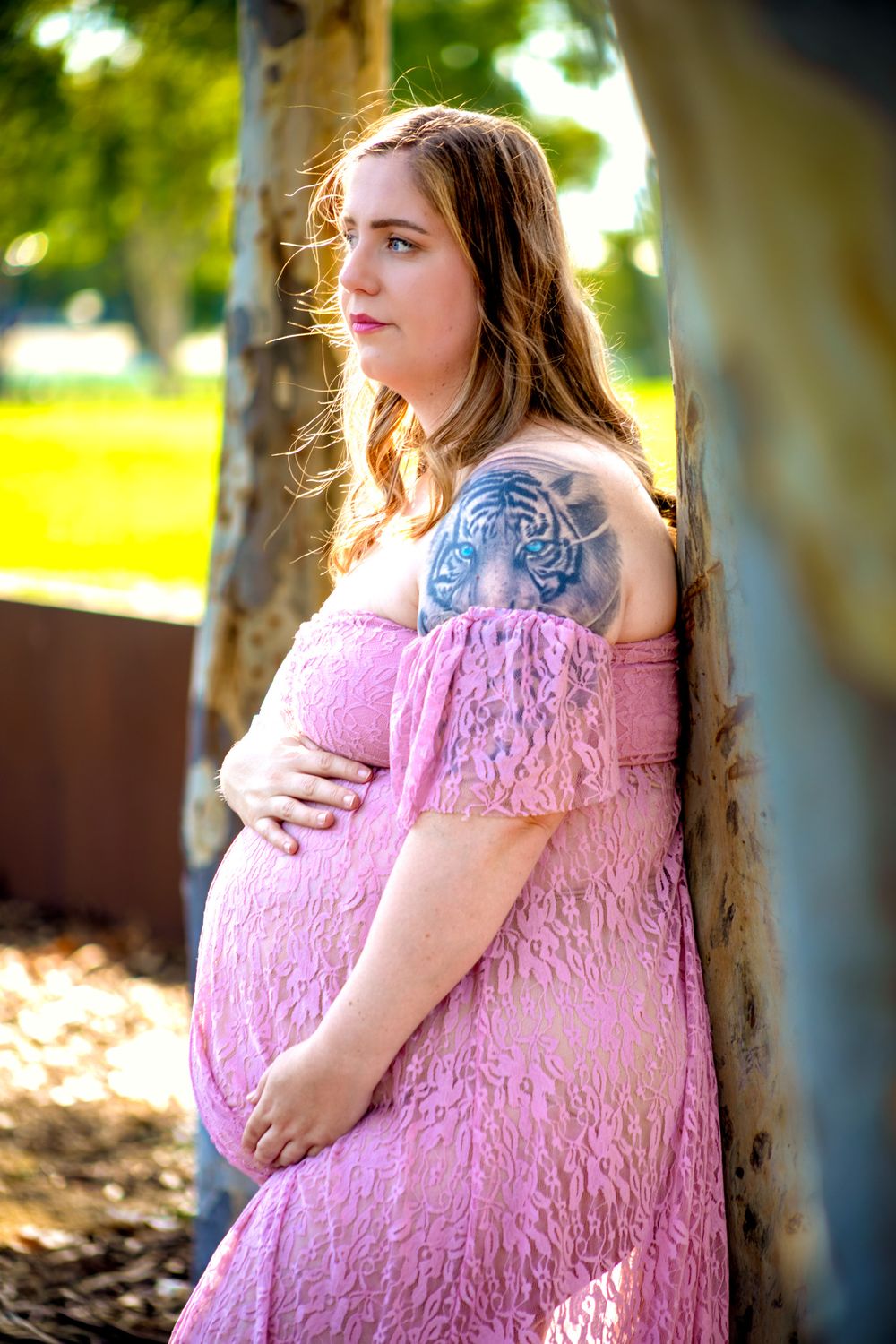 Pregnant woman in pink lace off-shoulder dress with tiger tattoo, posing outdoors in golden sunlight.