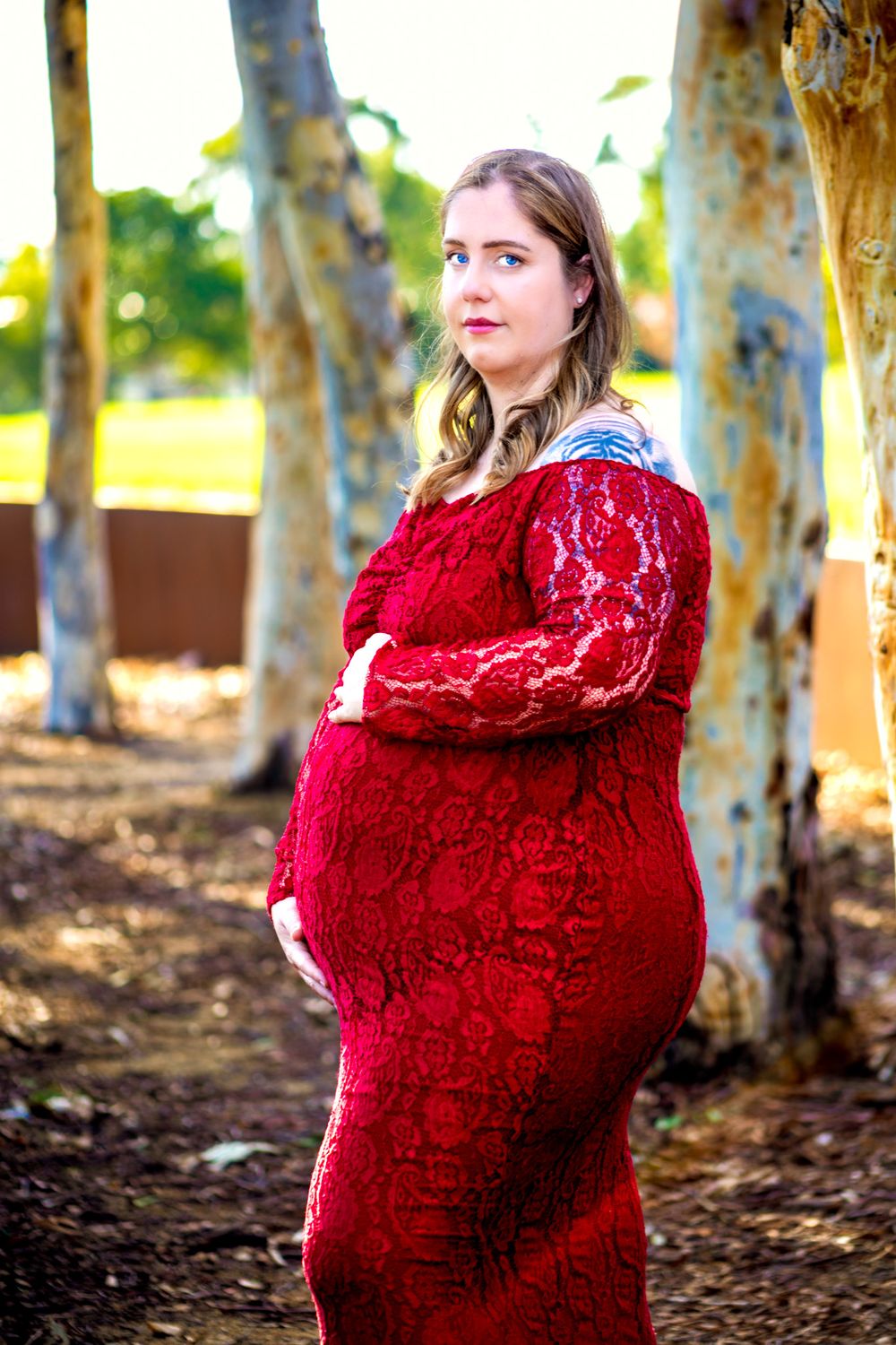 Pregnant woman in red lace dress cradling baby bump outdoors among trees during maternity photo session.