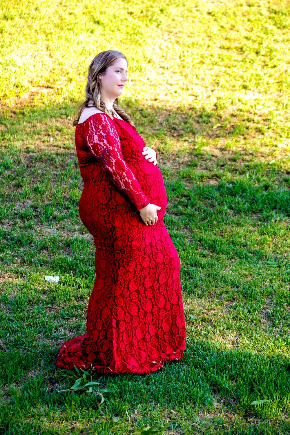 Pregnant woman in red lace gown standing outdoors on green grass, cradling her baby bump in a maternity photo.