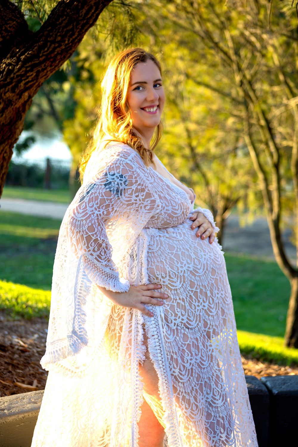 Pregnant woman in white lace dress holding baby bump, smiling outdoors in sunlit park with green trees.