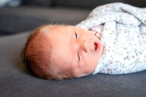 Baby wearing floral print sleepwear rests on dark grey couch with eyes closed.