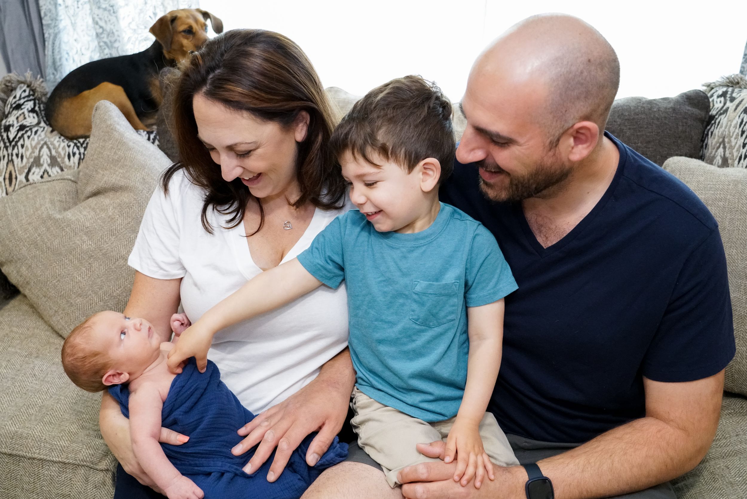 Family sits together on grey couch with dog overlooking a tender moment.