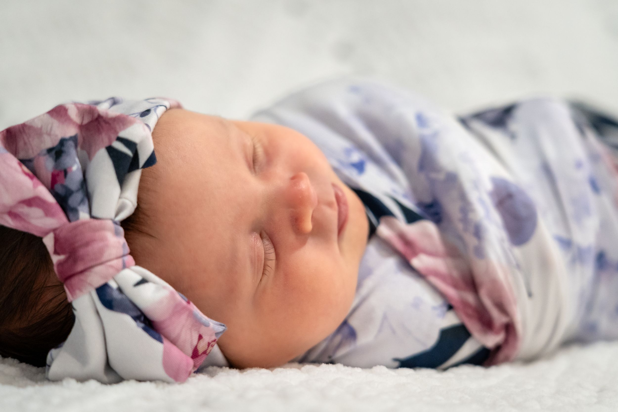 A sleeping newborn baby swaddled in floral print blanket rests peacefully on white bedding.