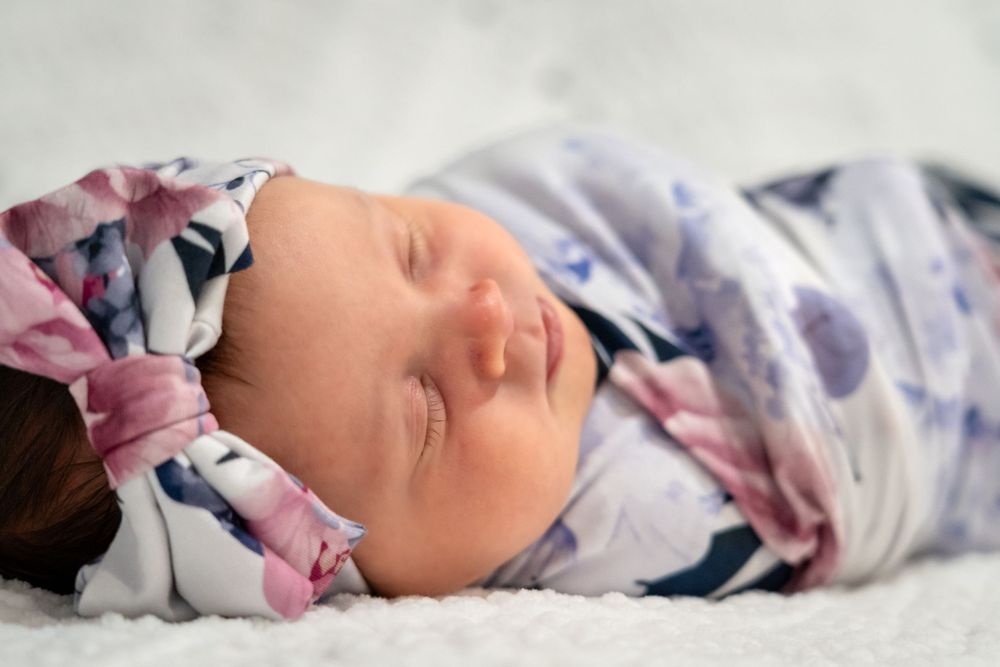 A sleeping newborn baby swaddled in floral print blanket rests peacefully on white bedding.