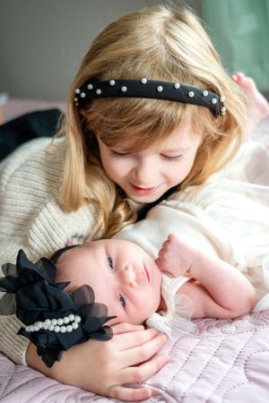 Sisters share sweet moment as older sibling cuddles with newborn on pink bedding.