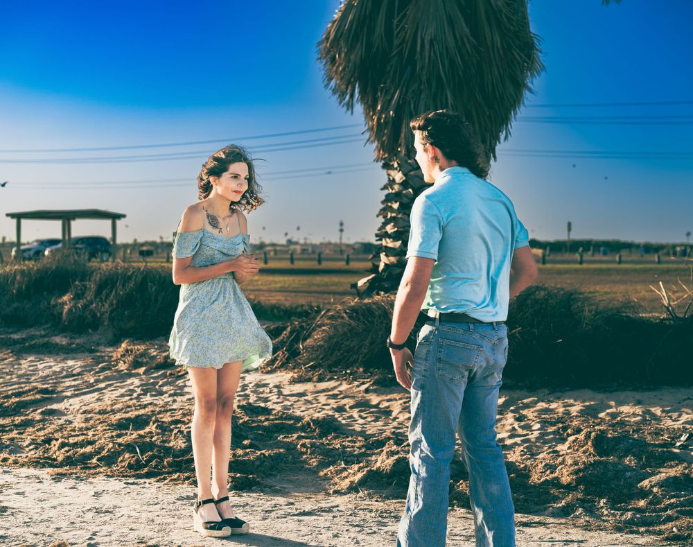 A couple shares a playful moment on a sandy beach with palm trees and blue sky in the background.