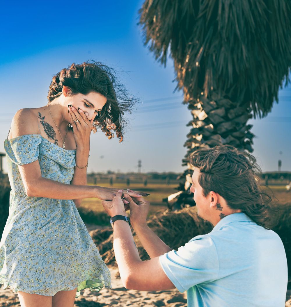 Under a palm tree and blue sky, a couple shares a joyful moment during a surprise proposal in a desert setting.