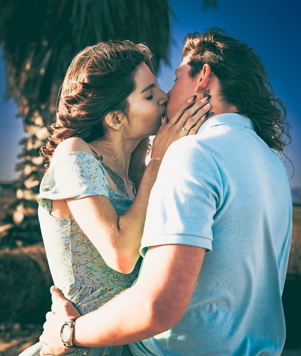 A romantic couple sharing a passionate kiss outdoors against a blue sky background with palm trees.