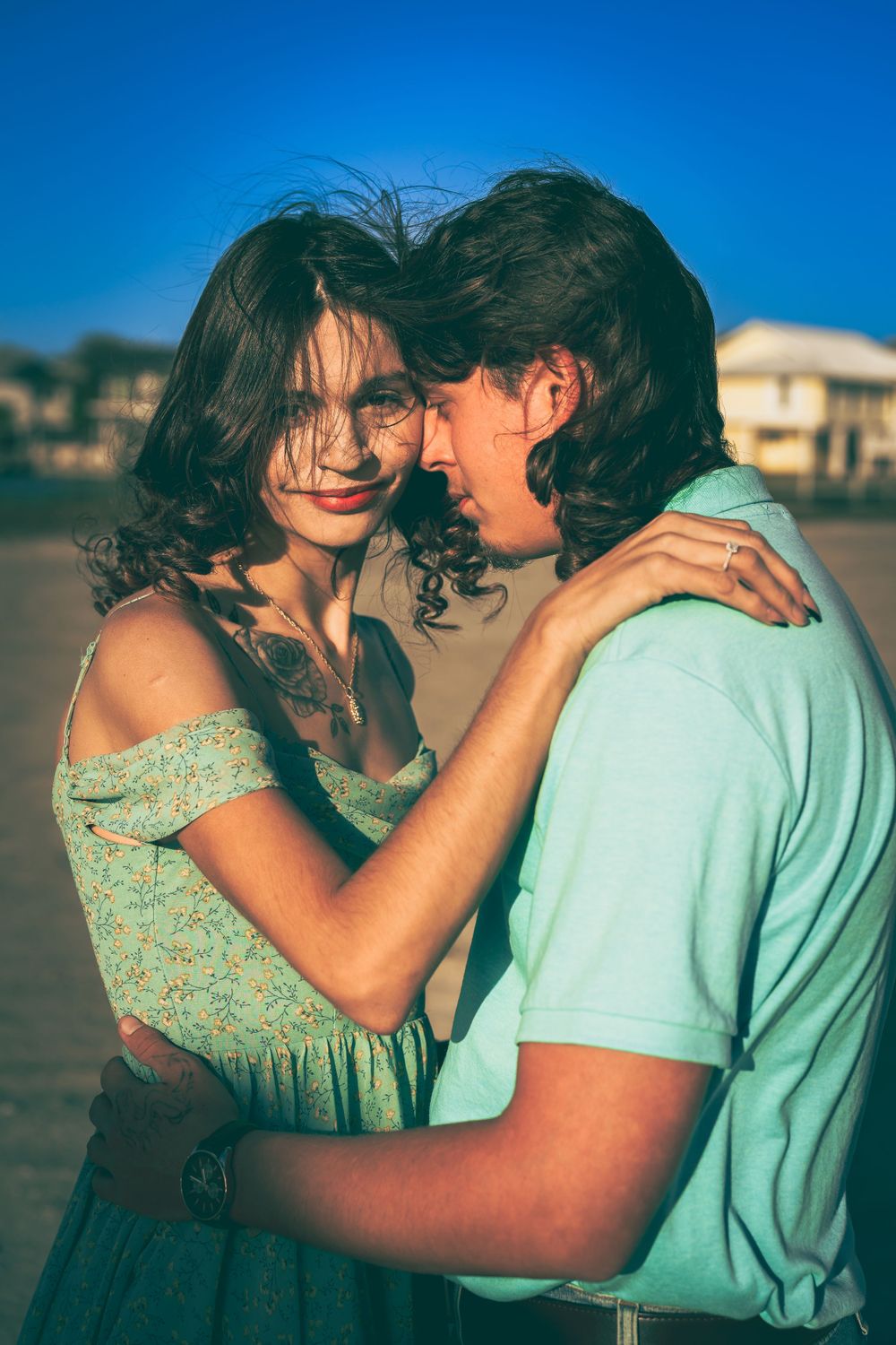 A romantic couple embraces and laughs together outdoors during golden hour, with a mint green shirt and floral dress.