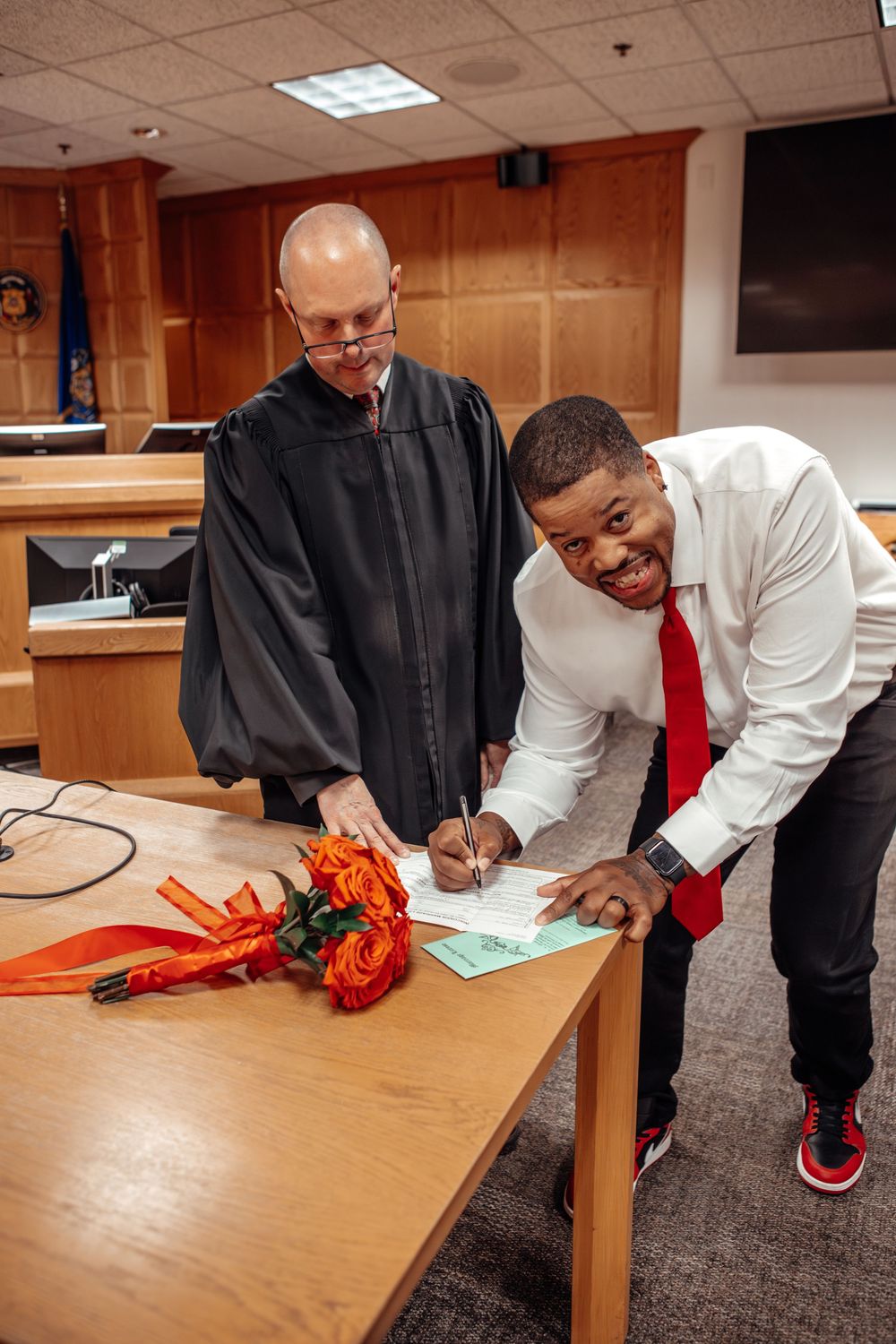 People in formal attire signing documents at a wooden table in an office setting.