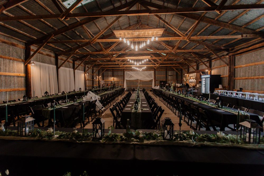 Long reception tables set up in a rustic barn venue with hanging chandeliers and industrial ceiling beams.