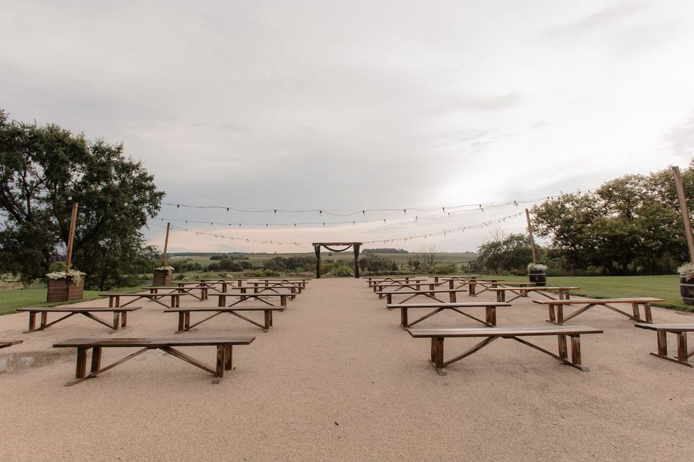 Outdoor wedding ceremony setup with rows of wooden benches under string lights at dusk.