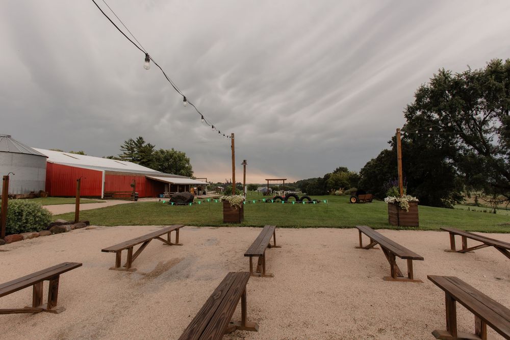 Rustic outdoor venue with wooden picnic benches and string lights under cloudy skies.