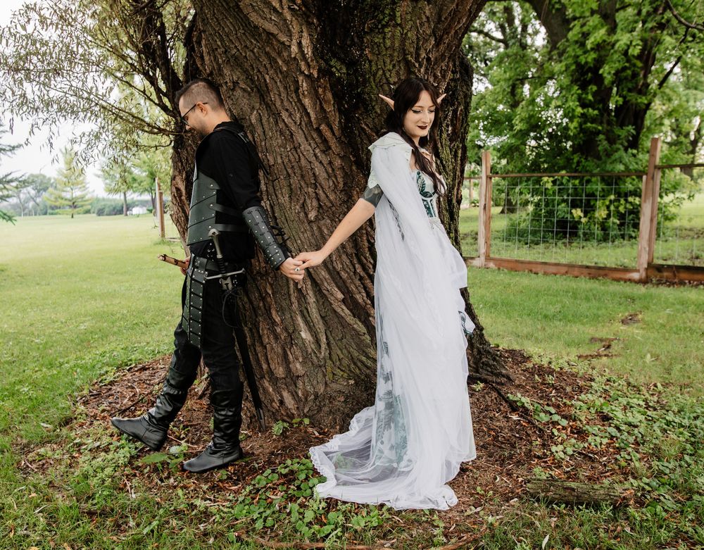 A couple poses back-to-back against a large tree trunk, with one in black attire and one in a flowing white dress.